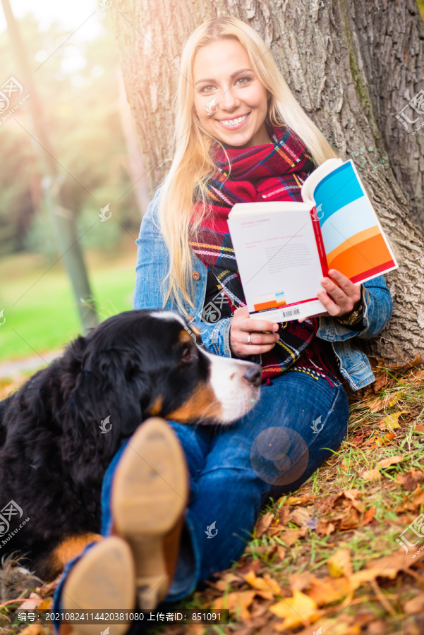 一位妇女和她的伯尔尼山狗在秋天公园里看书，坐在长满树叶的草地上