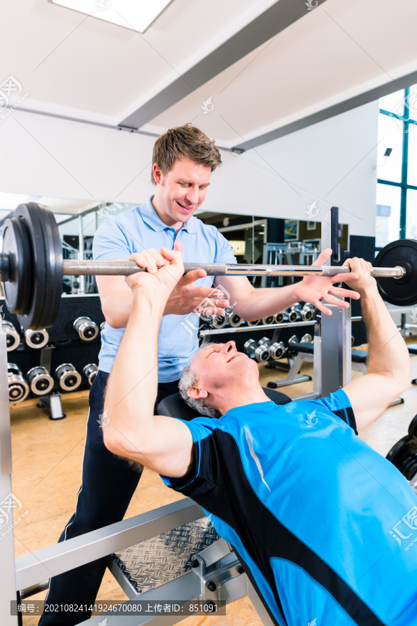 教练协助高级男子在健身房举重杠铃，以增加力量和健身