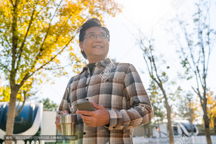 一个老年男人站在露营院子里拿着手机喝咖啡