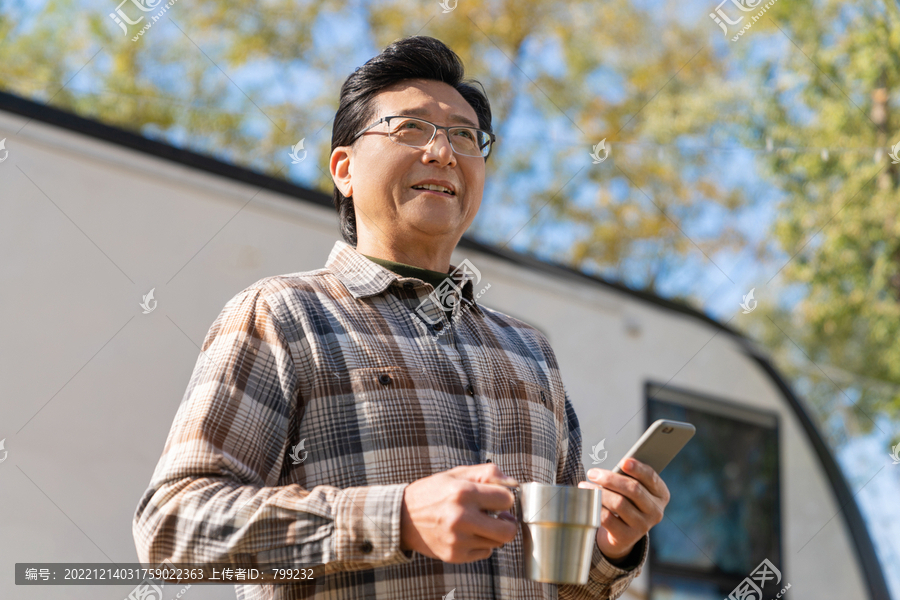 一个老年男人站在房车前拿着手机喝咖啡