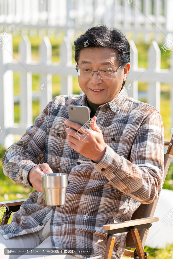 老年男人坐在露营院子里看着手机视频喝咖啡