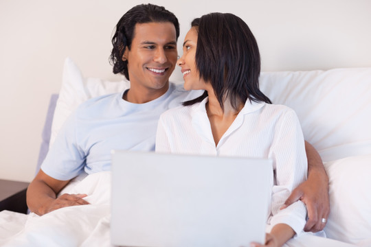 微笑的年轻夫妇坐在床上用电脑