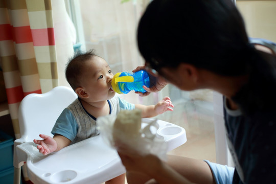 妈妈给坐在餐桌餐椅上儿子喂水喝