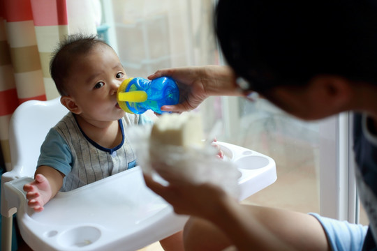 妈妈给坐在餐桌餐椅上儿子喂水喝