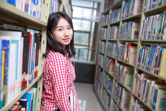 微笑的女学生靠在书柜上戴着耳机听歌