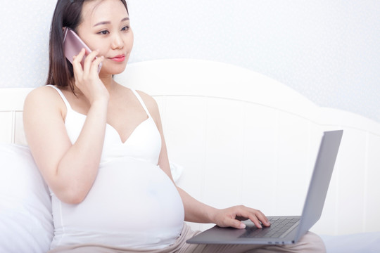 孕妇坐在床上使用笔记本电脑和移动设备