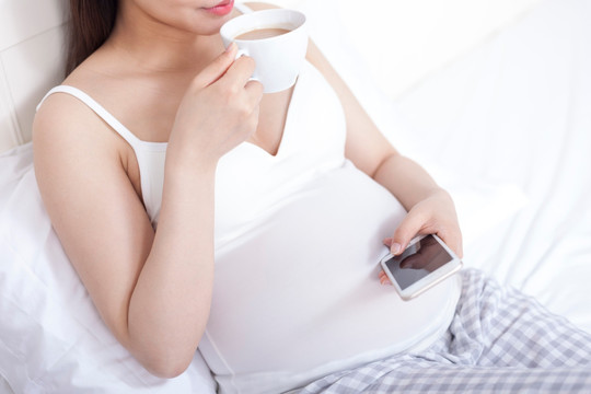 孕妇坐在床上一边使用手机一边喝着饮料