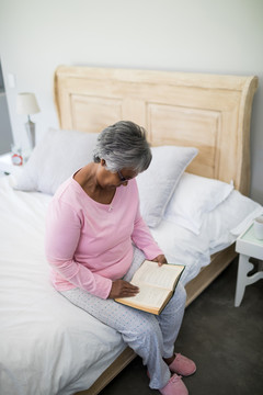 老年妇女坐在家里卧室的床上看书
