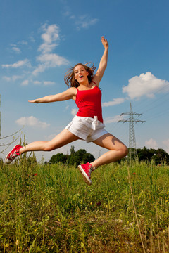 一位身穿红色t恤的妇女在蓝天下的夏日草地上跳得很高，背景是一根电线杆