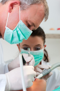 女患者带着牙医和助手在进行牙科治疗，戴着口罩和手套，牙医和助手用吸盘和钻头向她鞠躬