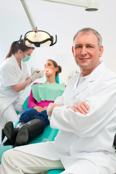 牙医在他的手术中看着观看者，背景是他的助手正在给一名女性患者治疗