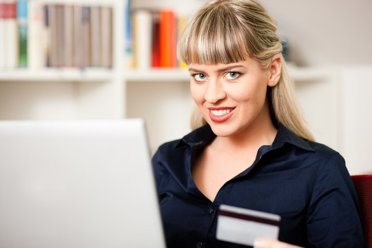一个女人拿着笔记本电脑坐在家客厅的书架前，在网上购物或做银行交易，背景是购物袋和信用卡