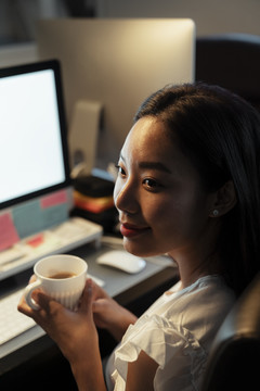 泰国亚裔女子晚上坐在办公室里端着一杯热咖啡的特写镜头。