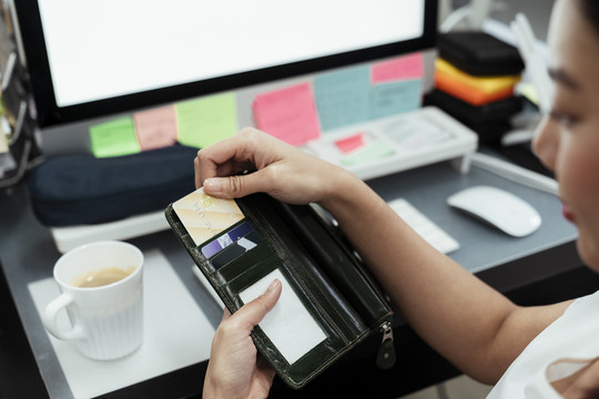 坐在电脑桌前，一位亚洲妇女从黑色钱包里掏出信用卡进行网上购物。