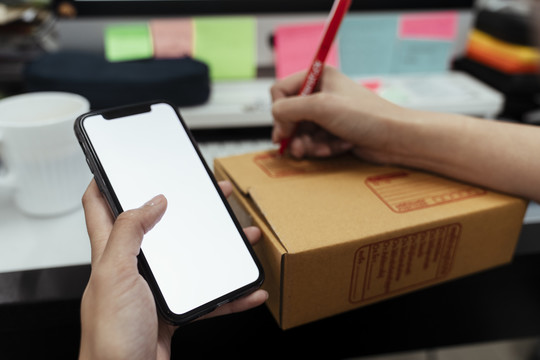 亚洲送货员的手在智能手机上阅读信息，然后用红笔在纸板箱上写下地址。