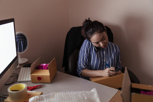 忙碌的亚泰妇女在纸板箱上写地址，和顾客通电话。复制计算机上的空间。