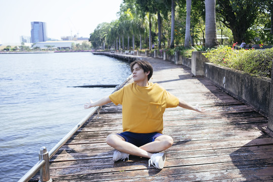 穿着黄色t恤和蓝色裤子的家伙坐在公园湖边的阳台上，张开双臂。