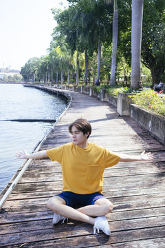 穿着黄色t恤和蓝色裤子的家伙坐在公园湖边的阳台上，伸开双臂，垂直。