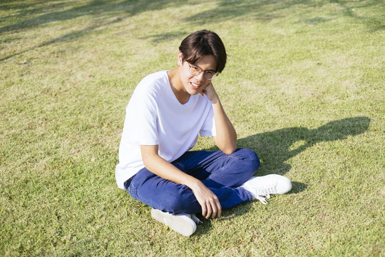 黑头发的家伙穿着白t恤，戴着眼镜，坐在草地上晒太阳。