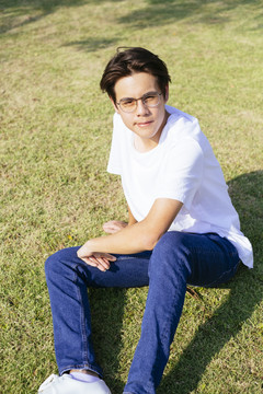 黑发小伙穿着白色t恤，戴着眼镜，坐在草地上，阳光直射，笔直。