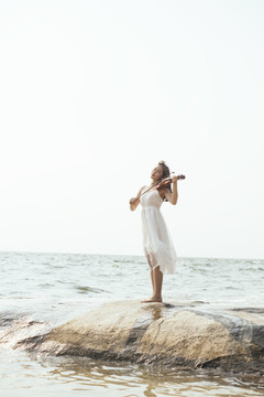 阳光明媚的日子里，一个穿着白衣服的女人在海边的岩石上拉小提琴。