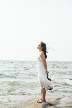 白裙女子站在海边的岩石上，闭上眼睛，背着小提琴感受着风。