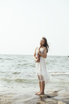 白裙女子闭上眼睛，怀里抱着小提琴，站在海边的岩石上，感受着风。