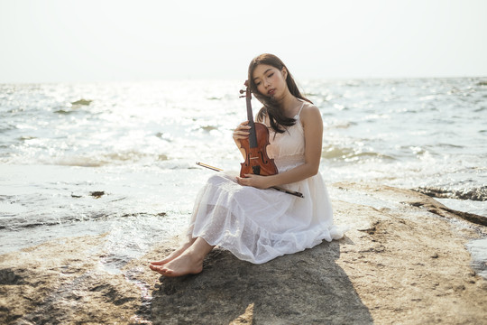 黑发白衣女子背着小提琴坐在海边的岩石上，享受阳光。