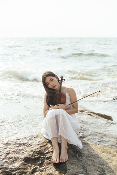 黑色长发，穿着白色连衣裙的女人，膝盖朝上，抱着小提琴坐在岩石上。