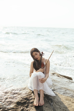黑色长发，穿着白色连衣裙的女人，膝盖朝上坐着，把小提琴放在岩石上。