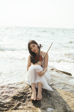 美丽的黑色长发女子穿着白色连衣裙，膝盖朝上，抱着小提琴坐在岩石上。