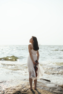 白裙女子站在岩石上面对太阳，把小提琴藏在背后的背影。