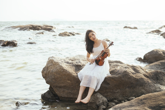 一位身穿白裙、赤脚的黑发女子手持小提琴坐在岩石上。