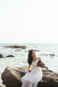黑发女子身穿白裙，赤脚坐在岩石上，拉着小提琴，闭上眼睛，以半海半天为背景。