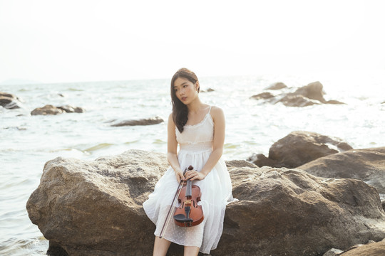 长发白衣女子从拉小提琴中解脱出来，坐在海边的岩石上。