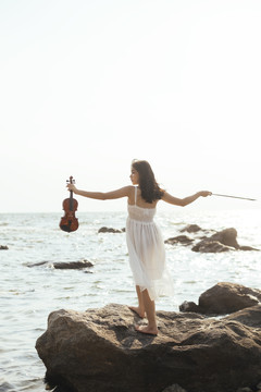 一位身穿白裙的黑发女子手持小提琴站在岩石上，张开双臂。