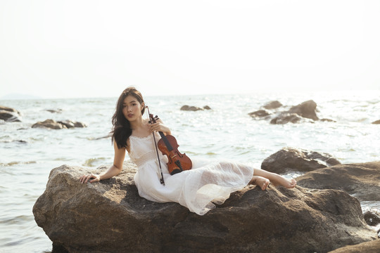 阳光明媚的日子里，一个黑色长发女人用右手肘躺在海边的岩石上，拉着小提琴。
