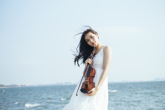 风大的日子里，站在海边，穿着白裙子的黑发女人把小提琴放在肚子上。