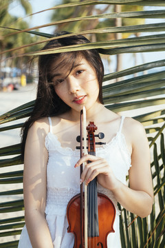 黑发女子身穿白裙，拉着小提琴站在椰子叶旁。