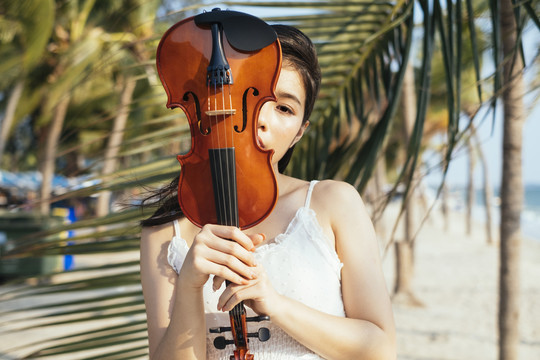 黑发白衣女子站在椰子叶旁拉小提琴掩面。