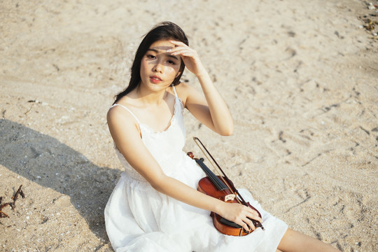 黑色长发小提琴手坐在沙滩上，拉着小提琴，举起手来保护眼睛不受阳光的伤害。