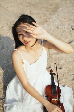 黑色长发小提琴手坐在沙滩上，拉着小提琴，举起手来保护眼睛不受阳光直射。