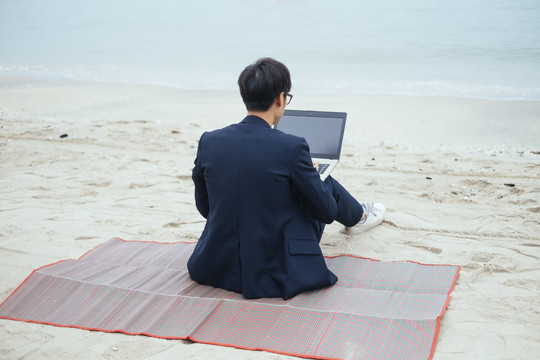 一个穿着深色西装的黑发男人坐在垫子上对着摄像机，手里拿着笔记本电脑在沙滩上工作。