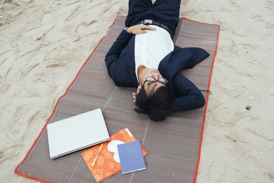 穿着深色西装的黑发男人躺在垫子上，手里拿着笔记本电脑和书躺在沙滩上。