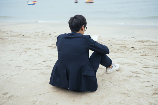 穿着深色西装的黑发男人坐在沙滩上对着镜头看书。