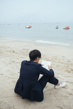 黑头发的家伙穿着深色西装，坐在沙滩上对着镜头，垂直地看书。
