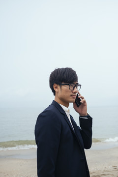 黑发黑衣人站在沙滩上，在朦胧的天空下接电话。
