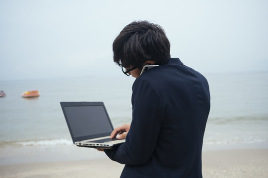 一个黑发黑衣人站在沙滩上和老板通电话，用笔记本电脑修改工作。