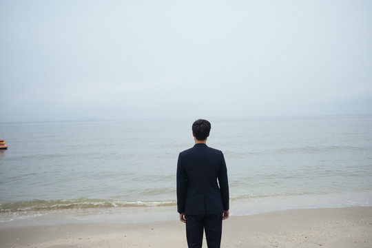 一个黑发男人，穿着深色西装，站在沙滩上，在阴暗的天空中看着大海。
