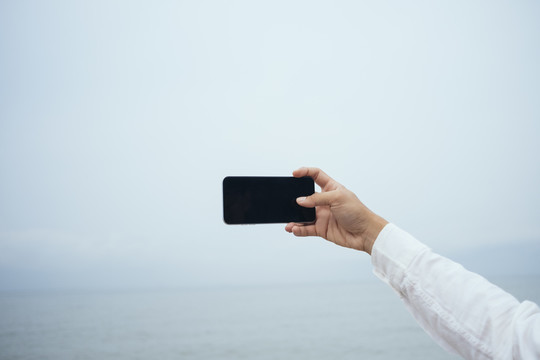 这名白衬衫男子在拍大海照片时，手里拿着一部智能手机的剪短图像。